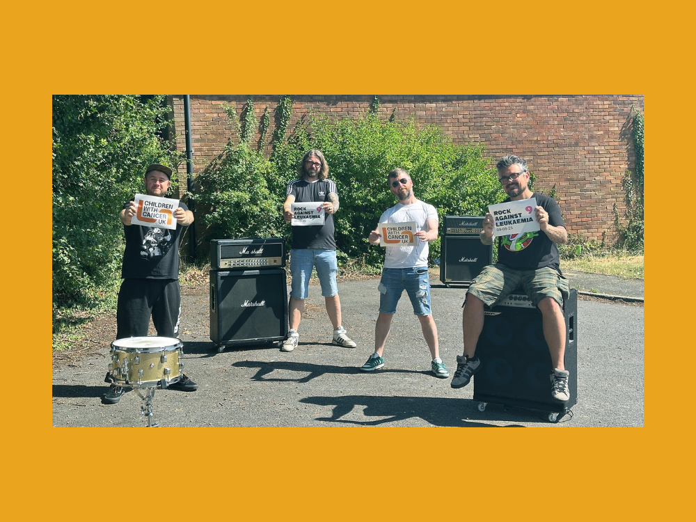 Four men posing outdoors in front of a brick wall and leafy greenery, holding signs that say “Children with Cancer UK” and “Rock Against Leukaemia 9.” Two men stand between Marshall amplifiers, one stands beside a snare drum, and one sits on a large speaker cabinet. All are casually dressed in T-shirts and shorts or jeans, with a sunny sky and shadows visible on the ground.
