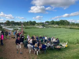 An open, green field and blue skies with lots of people sitting out on chairs with tables .